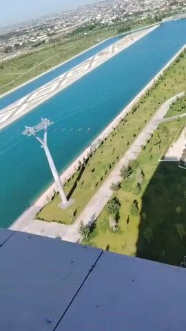 The video begins with an aerial view of a long, narrow, turquoise body of water with white lane markers, possibly a rowing course or canal. There are small red buoys marking the lanes and what appear to be a few small boats or kayaks in the water. A tall, white, pole-like structure with cables extending from it stands on the bank of the water, likely part of a zip line or similar attraction. Adjacent to the water is a grassy park area with scattered trees and paved walkways. Further in the distance, there is a city or town with buildings and roads. As the camera pans to the right, a modern, multi-story building with a green, angled roof comes into view. The building is surrounded by more parkland. The overall scene appears to be a well-maintained recreational or urban park area on a clear, sunny day. The video is shot from a high vantage point, possibly from a building or drone.