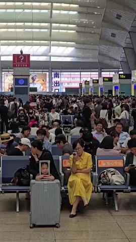 The video shows a crowded indoor waiting area, likely a train station. Many people are seated on blue benches, waiting. Some are looking at their phones, others are looking around, and some appear to be sleeping. In the foreground, a woman in a yellow dress is looking at her phone. Next to her, a young child is seated on a suitcase, holding a tablet. Luggage is visible around the seating area. The environment appears to be a large, open space with a high ceiling and fluorescent lighting. Signs with Chinese characters are visible in the background. The overall atmosphere is one of waiting and anticipation.