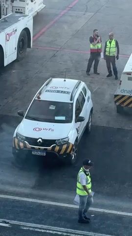 A high-angle, eye-level full shot captures a ground crew at an airport. Two men are standing near a white Renault Duster vehicle with "wfs" logos and an "ORB-4333" sticker on the roof. One man, wearing a red vest over a dark t-shirt and dark pants, is holding a clipboard and looking at the other man, who is wearing a yellow vest over a dark t-shirt and dark pants, looking away. A third man, also wearing a yellow vest, is walking from right to left, carrying a clipboard. The Duster has "wfs" logos on the side, a yellow and black striped bumper, and a license plate reading "TLE4J33." Another white vehicle with a yellow and black striped bumper and "dnata" text is visible on the right. The ground is dark gray with a pink line. The image appears to be taken from inside an airport terminal through a window.
