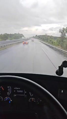 The video is filmed from the perspective of someone inside a truck, looking out the windshield. It is raining, and the road is wet, with water spraying up from the tires of passing vehicles. Several cars are visible on the road ahead, driving in the same direction as the truck. The sky is overcast and gray. The truck's dashboard is partially visible in the foreground.