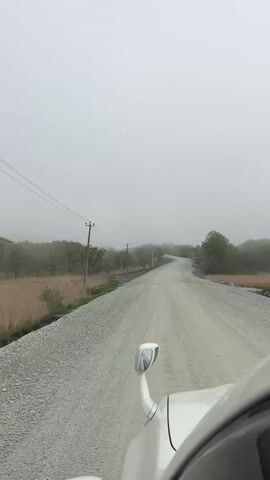 The video shows a forward-facing view from a car driving on a gravel road. The road is surrounded by trees and tall grass, and power lines run along one side. Fog is present, obscuring the distance. The car appears to be moving at a moderate speed. The side mirror of the car is visible in the lower right corner of the frame. The overall impression is one of a remote, possibly rural, setting.