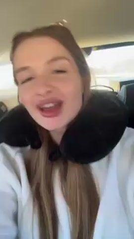 A young woman wearing a white sweatshirt and a black travel pillow is recording a video of herself while in a car. She has long, straight brown hair, fair skin, and is wearing lip gloss. She makes various expressions with her mouth while music plays. The interior of the car is visible in the background.