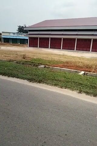 The video shows a view from a moving vehicle, possibly a car or motorcycle, traveling alongside a road. On the left side of the road, there is a building with a red and white facade, likely a warehouse or industrial building. Further down the road, there is a smaller blue and white building with a sign that appears to be written in Arabic script. There are also several trees, including a palm tree, and some grass and dirt areas. The sky is overcast, and the overall scene appears to be rural or semi-rural.