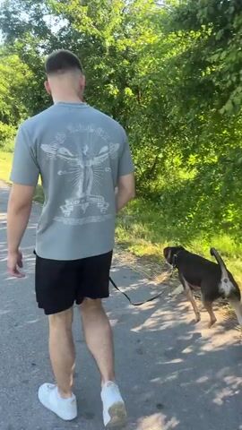 A man and his dog walk down a paved path surrounded by trees. The man is wearing a grey t-shirt and black shorts, and the dog is brown and black. The sun is shining and the sky is blue. The path appears to be a walking or biking trail.