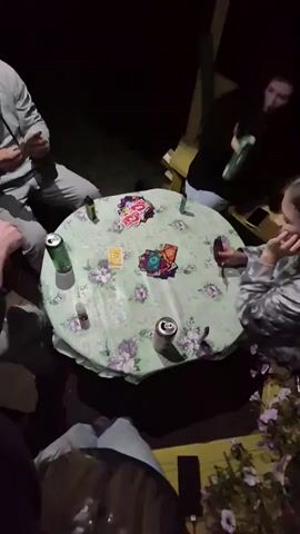 The video shows a group of people sitting around a round table at night. The table is covered with a light green tablecloth with a floral pattern of purple and white flowers. On the table, there is a deck of cards, some of which are spread out in the center. There are also several cans of what appear to be beverages and a smartphone. The people are casually dressed and appear to be engaged in a game. The lighting is dim, suggesting an outdoor setting at night, possibly a patio or deck, with some artificial light illuminating the table. One person on the left is holding cards in their hand and looking at them. Another person on the right is also holding cards. The overall atmosphere seems relaxed and social.