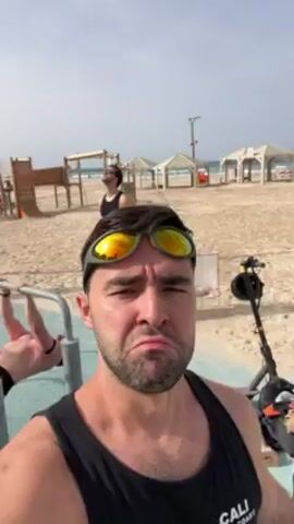 A man is filming himself at a beach during the day. He is wearing a black tank top and green framed mirrored sunglasses. He is making a call-me sign with his hand. There are people, sand, and cabanas in the background.