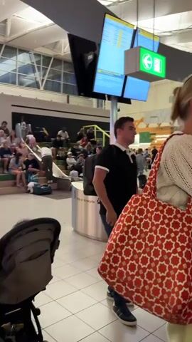 The video shows a busy airport terminal. People are seen sitting on benches, waiting, and walking with luggage. In the foreground, a person is looking at their phone while sitting on a green bench. To the right, a store labeled "PERFUMES & COSMETICS" is visible. There are also displays of products and signs within the store. In the background, more people and shops are seen, and there are signs indicating "LOUNGE 4". The overall environment is bright and modern, typical of an airport setting, with a high ceiling and glass structures. The footage is slightly blurry due to movement.