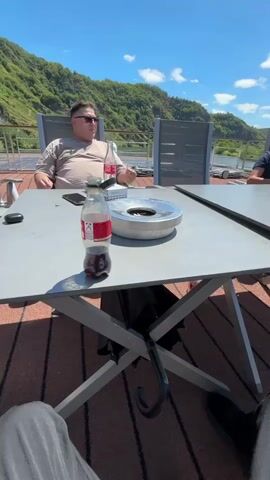 A group of men are sitting around a table outdoors on a sunny day. The table is set up on a deck or patio, with chairs around it. There are several bottles of Coca-Cola on the table, as well as an ashtray. One man is smoking and looking away from the camera. Another man is holding a bottle of Coca-Cola and looking at his phone. A third man is standing and appears to be preparing something on the table. In the background, there are trees, hills, and a body of water. The sky is blue with some clouds.