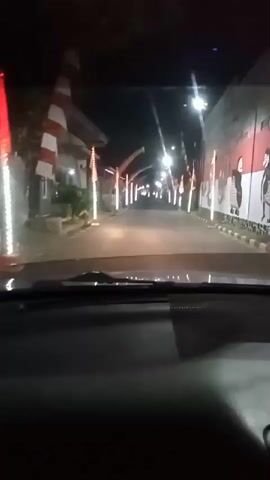 The video shows a view from inside a vehicle driving down a road at night. The road is lined with lights and decorations, including red and white flags. On the right side of the road, there is a wall with murals painted on it, depicting figures in what appears to be traditional clothing. The murals are in red and white, matching the colors of the flags. The overall scene suggests a festive or celebratory atmosphere, possibly related to a national holiday. The video is shaky, indicating it was likely recorded by someone holding a phone or camera while the vehicle was in motion.