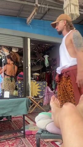 The video shows a man standing in a market setting, wearing a white tank top, a baseball cap, and leopard print pants. He has a light brown garment tied around his waist and a tattoo on his left arm. He is looking toward the market stall, where a woman is arranging items on a table. Another man sits at the table, wearing a floral shirt and talking on a cell phone. The market stall is filled with various items, including clothing, accessories, and decorations. The floor is covered with a patterned rug. The camera is positioned close to the ground, showing a pair of bare feet resting on cushions in the foreground.