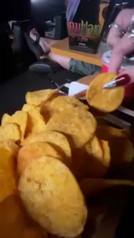 A person with long brown hair and bangs sits in what appears to be a movie theater. The person dips a large corn chip into a white sauce and then eats it. The shot then returns to the pile of corn chips.