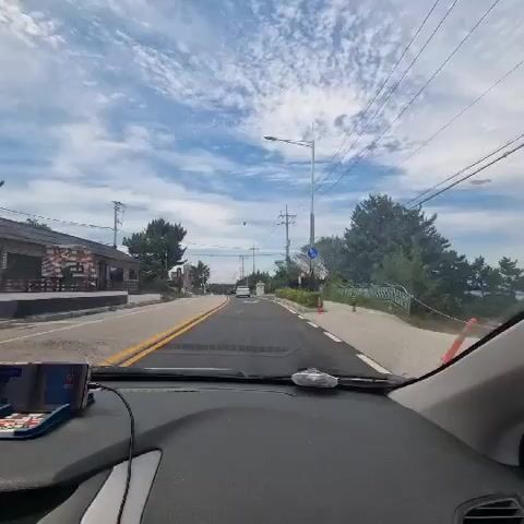 This video is a first-person perspective from inside a car, looking out the windshield as the car drives down a paved road. The sky is blue with scattered white clouds. There are trees and power lines visible along the sides of the road. On the left side of the road, there are buildings and what appears to be a restaurant or shop. On the right side of the road, there is a sidewalk and more trees. The road is marked with yellow and white lines. There is a speed limit sign and a blue directional sign visible on the right side of the road. A mobile phone is mounted on the dashboard.