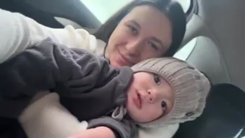 A woman is holding a baby in her lap in the back seat of a car. The woman is fair-skinned with long, dark hair and is wearing a white sweater. Her eyes are closed and she is smiling. The baby is fair-skinned with a gray hat and a gray jacket. The baby is looking at the camera. The car is moving, as evidenced by the background blurring outside the window. The interior of the car is visible, including the door panel and window controls. A low hum or drone sound is audible.
