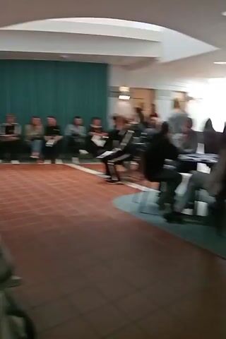 The video appears to be filmed in a public indoor space, possibly a community center or educational institution in Kuopio, Finland. The scene features several groups of people, predominantly young adults, engaged in various activities. In the foreground, a person with blonde hair and a plaid shirt is partially visible from behind, facing away from the camera. Further back, a group of individuals, likely students, are seated in a semi-circle, facing a teal-colored wall, suggesting a presentation or discussion is taking place. They are casually dressed, and their demeanor suggests attentiveness.

To the left and right of this central group, other individuals and smaller groups are seated at tables or on chairs. Some appear to be engaged in conversations, while others are observing the proceedings. The lighting is bright and diffused, indicating it's daytime. The architecture suggests a modern building with high ceilings and ample natural light, possibly from skylights. The flooring is tiled, and the walls are a mix of painted surfaces and what looks like acoustic paneling.

There is a visible sign that says "WC" indicating a restroom facility. The overall atmosphere seems relaxed and informal, with no overt signs of distress or urgency. The people are dressed in casual, everyday clothing, and there are no indications of specific weather conditions as the scene is entirely indoors. The presence of multiple people gathered suggests a planned event, a class, a meeting, or a social gathering. The time of day is likely during daylight hours, given the bright indoor lighting.