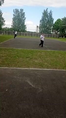 The video shows two young people in uniform engaged in a physical activity on a sports court. One person is running across the court, while the other is walking in the opposite direction, holding an object. They appear to be part of a group of people gathered beyond the court, visible along the fence. The environment includes a grassy field adjacent to the court, with trees surrounding the area. The sky above is partially cloudy.