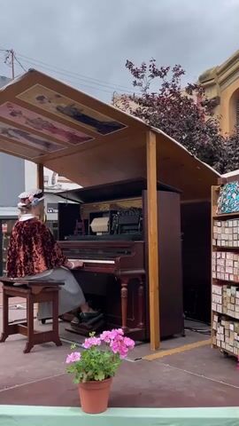 A woman wearing a burgundy cape, grey skirt, and decorated hat sits at a dark brown, antique-style piano outside on a red stage. The piano is housed in a wooden structure with paintings on the underside of its roof. She is operating the piano, and rolls of paper are visible inside it. Beside the piano, there is a rack of music rolls. In front of her, there is a pot of pink geraniums. In the background, a street scene with shops and buildings is visible under a cloudy sky.
