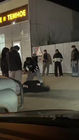The video captures a nighttime scene in Kizner, Russia, where a group of approximately 8-10 young men are gathered outdoors on a paved surface. A violent physical altercation is taking place at the center of the group. One individual, dressed in dark clothing, is on the ground, seemingly being held down or assaulted by another person. The aggressor is wearing a dark hoodie with the letters "CCCP" (SSSR) prominently displayed on the back, and grey pants.

Several other men stand around, observing the ongoing incident without intervening. One person in the background is seen pointing towards the altercation. The scene is illuminated by artificial light, primarily from a brightly lit sign on an adjacent building. The sign clearly displays the Russian words "СВЕТЛОЕ И ТЕМНОЕ" (SVETLOE I TEMNOE), which translates to "LIGHT AND DARK" (commonly used for types of beverages).

The footage appears to be filmed from inside a vehicle, with a portion of a white car visible in the immediate foreground. The overall atmosphere of the scene is tense and depicts an act of aggression in a public space.