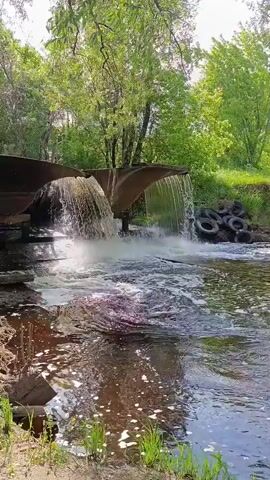 This video displays a waterfall created by a man-made structure, possibly a dam or spillway, made of metal and stone. The water cascades down into a river, creating a foamy and turbulent flow. The surrounding environment is lush and green, with trees and vegetation evident along the riverbanks. In the background, on the right side of the frame, a pile of old tires is visible, suggesting a potential environmental concern. The camera pans slowly across the scene, capturing the movement of the water and the natural beauty of the area. The overall impression is one of a somewhat neglected but still picturesque natural setting.