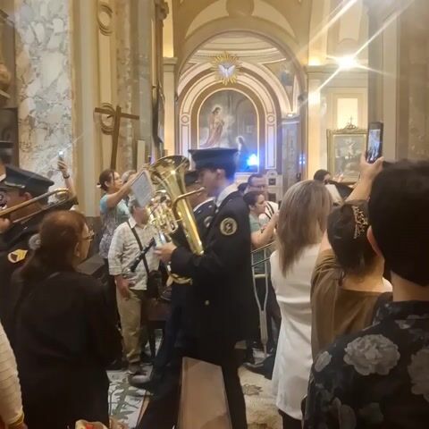 Inside an ornate church, a band of men in uniform plays brass instruments, surrounded by a crowd of people. Many are holding up their phones, recording the performance. The church has tall arches and pillars, with religious paintings and sculptures visible in the background. The atmosphere is lively and festive.