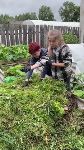 Two children, a boy with red hair and a girl with blonde hair, are working in a garden. They are both wearing black gloves and are handling a large pile of cut grass and plants. The boy is bent over the pile, while the girl is kneeling beside it, also working with the material. The background shows a wooden fence, some plants, and what appears to be a greenhouse. The overall scene suggests they are engaged in gardening or composting activities.