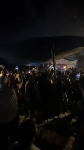 The video shows a crowded outdoor gathering at night, likely a party or concert. A large group of people are packed together under a makeshift canopy or tent. Many individuals are holding up their phones, seemingly recording or taking pictures. The lighting is dim, with some illumination coming from stage lights or the phones themselves. The atmosphere appears energetic, with people raising their hands and seemingly engaged in the event. The ground looks sandy or dusty.