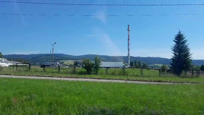The video shows a view of a rural landscape under a clear blue sky. In the foreground, there is a large area of green grass. A metal fence runs horizontally across the middle of the frame, separating the grassy area from the background. Behind the fence, there are several industrial structures, including a tall, slender tower with red and white markings and what appears to be a building or factory. Further in the background, rolling hills covered in trees are visible. There are also some electrical poles and wires scattered throughout the scene. The overall impression is one of an industrial facility situated within a natural, possibly mountainous, environment.