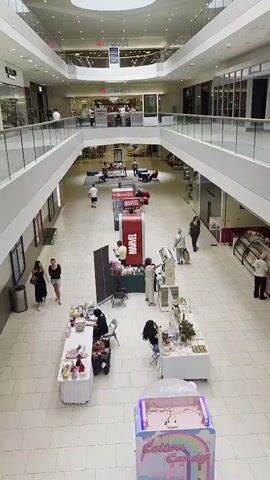 The video shows a multi-level shopping mall interior. On the ground floor, several pop-up booths are set up along the walkway. One booth displays products with Marvel branding. Another booth appears to be selling handmade items like jewelry and skincare. A cotton candy machine is visible in the foreground. People are seen browsing the booths and walking through the mall. The upper level features shops and balconies overlooking the ground floor.