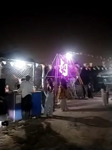 A video clip at night captures an outdoor gathering with a temporary setup. In the foreground, a blue and white counter is visible, with a person in a white shirt and dark pants standing behind it. To the left of the counter, a chain-link fence encloses a space. Several people, appearing to be young adults, are gathered in front of and around the counter. One person, a female wearing light-colored pants and a dark top, stands close to the counter. To her right, another female in a patterned top and light pants is also present. In the background, a large structure, possibly a ride or a stage, is illuminated with purple and white lights. Decorative triangular flags are strung across the top of the scene. The ground appears to be unpaved, with loose dirt and debris. The overall atmosphere suggests a nighttime event or fair.
