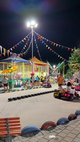 The video depicts a nighttime scene at what appears to be an amusement park or fairground. Colorful pennant flags are strung across the frame, illuminated by overhead lights and the glow from various attractions. In the foreground, a go-kart track is visible, with several children and adults riding in small electric cars. The track is bordered by tires, and some of the karts have flashing lights. To the left, there is a play structure with slides and colorful panels, with people gathered around it. Further in the background, a bungee trampoline attraction is in operation, with individuals being launched into the air. People are seen sitting on benches, walking around, and interacting with the attractions. The overall atmosphere is lively and festive.