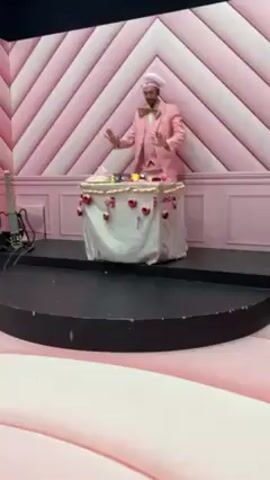 The video shows a DJ standing behind a table covered with a white cloth, performing on a small, black stage. The DJ is wearing a pink suit with a bow tie and a chef's hat. The table is decorated with red ornaments and musical notes. Behind the stage is a pink and white striped wall with paneling. To the left of the DJ, an electric guitar rests on a stand. The base of the platform is light pink with darker pink accents. The DJ is actively using DJ equipment on the table, gesturing to the side, and adjusting controls. The music playing has a techno beat.