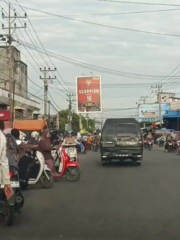 This is a medium shot, street-level, wide-angle video of a busy road with moderate traffic, primarily consisting of motorcycles and a few cars. The sky is overcast with a pale grey hue. On the left side of the road, there are buildings with storefronts, and power lines crisscross overhead. A prominent billboard in the center background advertises "SCORPION 16" with a price displayed below. Several people are riding motorcycles, most wearing helmets and some wearing headscarves. One person on a white scooter is looking towards the camera. A red scooter is in the foreground, with a person wearing a brown headscarf and a pink shirt riding it. A black van is moving away from the camera in the center of the road, and other motorcycles are following behind it. The road surface appears to be asphalt, and there are white lane markings visible. The overall impression is a typical urban street scene during the day, likely in Southeast Asia due to the prevalence of motorcycles and the style of clothing.