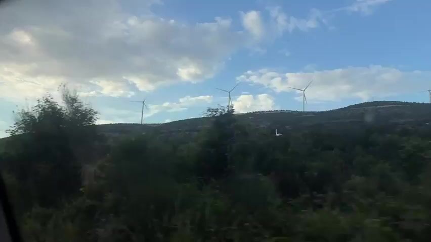 The video shows a car driving down a road, with the camera positioned inside the car.  Outside the car, there are several wind turbines visible on top of a green, tree-covered hill against a partly cloudy sky.  The camera angle shifts briefly to focus on a single turbine. The view then moves to show the car's interior, focusing on the dashboard, steering wheel, and the back of a person's head. Other cars are visible on the road ahead. The environment appears to be rural, with a winding road.