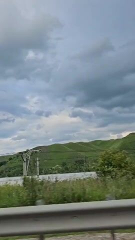 The video shows a view from a moving vehicle traveling alongside a body of water and green hills under a cloudy sky. In the foreground, a silver guardrail is visible. Beyond the guardrail, tall green grass and wildflowers line the roadside. Further back, a river or large creek flows, bordered by trees and more vegetation. The rolling hills are covered in lush green grass, with scattered trees and some white structures or patches of lighter ground visible on their slopes. Utility poles and wires are present along the landscape. In the mid-ground, several white buildings with dark roofs are situated. The sky is filled with heavy, gray clouds, suggesting overcast or potentially stormy weather.
