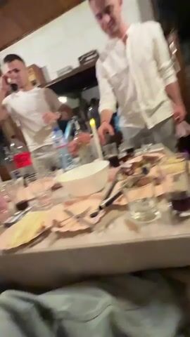 The video shows a group of people gathered around a table in a room with a rustic interior. Two men are prominent in the foreground: one is on a phone call while holding a glass, and the other is standing near the table, holding a glass of dark liquid. The table is cluttered with used plates, cutlery, glasses, bottles of water and soft drinks, and food remnants. A white bowl and a lit candle are also on the table. The atmosphere suggests a gathering or meal has taken place. The camera angle is low, appearing to be at a seated or reclined position.