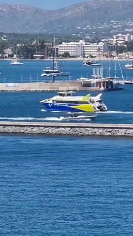 A blue and yellow catamaran speeds across the bright blue water, leaving a white wake. The boat is moving from left to right in the frame. In the background, a stone breakwater runs horizontally across the middle of the shot. Beyond the breakwater, there are several other boats, including sailboats and smaller vessels, moored in a harbor. Further in the distance, a coastal town with white buildings and some trees is visible on a hillside. The sky is clear and sunny.