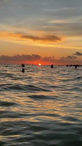 The video shows a body of water, likely the ocean, with numerous people swimming and wading in it. The sun is setting, casting a warm orange and yellow glow across the sky and reflecting on the water's surface. Clouds are present in the sky, adding texture to the sunset. The waves are moderate, creating ripples and patterns on the water. The overall scene depicts a lively and scenic beach environment during sunset.