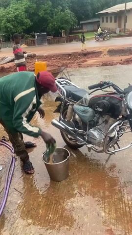 A person wearing a green jacket and a red cap is washing a black motorcycle. They are using a cloth to wipe down the exhaust pipe of the motorcycle. A metal bucket filled with water is on the ground next to the motorcycle. There is a purple hose on the ground to the left of the person. In the background, there are other people and motorcycles, and the area appears to be a construction site with dirt and puddles of water.