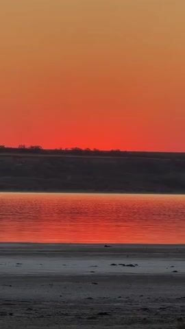 A vertical video displays a serene landscape at dusk. The top portion is a gradient of orange and red sky, with a dark, silhouetted horizon line of hills or land. Below the horizon is a calm body of water, reflecting the vibrant colors of the sky, creating a shimmering red and orange surface. The foreground is a beach or shore with a sandy and rocky texture, appearing in shades of gray and white. The water is still, and the overall scene conveys a sense of peacefulness and natural beauty.