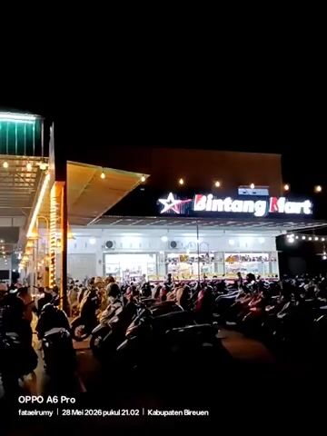 At night, the camera slowly pans across an outdoor scene in front of what appears to be a large, well-lit food court or market. Many people can be seen walking around and patronizing the various establishments within. The structures have a metal and corrugated roof with festive lights strung throughout. Numerous motorcycles are parked in the foreground, some closely packed together. A tree is decorated with string lights. The scene is captured with an OPPO A6 Pro, with a timestamp indicating May 28, 2026, at 21:02 in Kabupaten Bireuen.