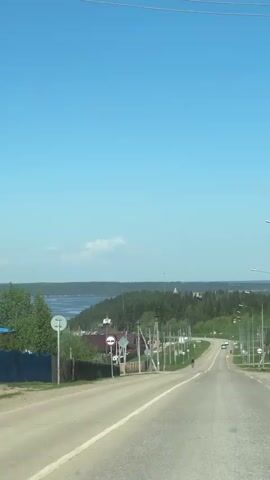 The video shows a drive through a rural area, starting on a paved road lined with utility poles and signs, passing houses with diverse roof colors towards a distant view of a wide body of water bordered by a dense forest under a bright blue sky with scattered clouds. A cyclist can be seen riding down the road in the same direction as the vehicle filming.