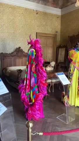 This is a video of an indoor display of colorful, feathered garments on mannequins. The room appears to be a museum or exhibition space, with ornate wooden furniture and patterned wallpaper. The lighting is dim and artificial. There are no people visible in the video. The camera pans across the garments, focusing on their vibrant colors and elaborate designs. The overall impression is one of opulence and extravagance.