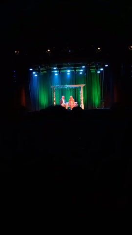 The video shows a stage performance from the perspective of an audience member in a darkened theater. The stage is set with a backdrop of green curtains and illuminated by blue and white stage lights.  A white decorative archway, adorned with greenery, frames a bench where two people are seated. A man in a light-colored shirt and darker jacket sits to the left, while a woman in a peach-colored dress sits to the right. The people appear to be engaged in a conversation.