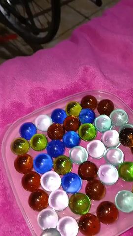 A hand is seen interacting with a collection of colorful glass marbles. The marbles are arranged in a clear plastic container, which is placed on a pink fuzzy surface. The marbles vary in color, including blue, green, clear, and brown. The hand reaches in and picks up a blue marble, which is then held close to the camera. The background is slightly blurred, but appears to be a room with some furniture. The lighting is bright, causing some glare on the marbles.