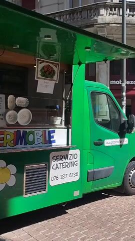 A bright green food truck, identified as "PIZZOCCHERI MOBILE," is parked outdoors on what appears to be a paved street or market area during the day. The camera pans slowly from right to left, revealing the side of the truck.

The truck features several notable decorations and signs. A large heart graphic is split into the Swiss flag (white cross on red) and the Italian flag (green, white, red vertical stripes), suggesting a connection between the two countries. Below this, the phrase "il piatto del gemellaggio" (the twinning dish) is written in a decorative script. Further down, a white flower decal has "100% vegetarian" written inside it.

A service window is open, revealing menu boards and food displays inside. Text on the menu boards includes "PIZZOCCHERI" with prices listed (Fz. 10.-- / Fz. 15.-- / Fz. 20.--), and "VASCHETTE D'ASPORTO" (takeaway containers). Another sign reads "NOI CONTANTE" with prices. There's also a QR code visible for digital payments or information. A display shows a picture of "Pizzoccheri," a traditional pasta dish.

Towards the rear of the truck, a sign clearly states "SERVIZIO CATERING" and provides a phone number: "076 337 6736." An "ORDER NOW" sticker with "smood.ch" and app store logos is also visible, indicating online delivery options. The truck's bright green color, clear daylight, and specific signage suggest it is operational and ready to serve customers in a European, likely Swiss, setting.