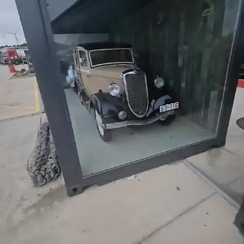 A short video shows a glass display case containing three vintage vehicles. First is a black and tan vintage car. Behind that car is a silver Vespa scooter. Reflected in the glass is a tan vintage car parked outside the display case. The display case is sitting in a parking lot. In the background, there are trucks, wind turbines, and some low-lying industrial buildings.