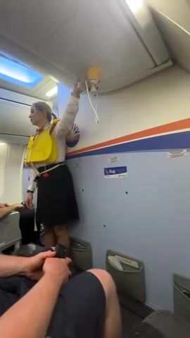 In an airplane cabin, a flight attendant is demonstrating how to use the oxygen masks in the event of a loss of cabin pressure. She is wearing a yellow life vest over her white shirt and a black skirt. With her left hand, she holds the yellow oxygen mask to the ceiling. The mask is connected to a long, thin, clear tube. Her right arm is raised to assist. The cabin walls are light blue with a horizontal stripe of red, white, and blue. A row of oxygen masks is recessed above her. A passenger in the foreground is holding a remote in his hands and watching the demonstration. Another passenger is sitting in the row in front of him. A small sign labeled "X-LUG" is on the wall beside the flight attendant.