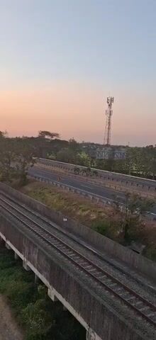 The video pans across a landscape at sunset. In the foreground, there are train tracks running parallel to a highway. Cars and trucks can be seen on the highway. In the background, there is a cell tower, trees, and fields. The sky is a gradient of orange and pink, with the sun partially obscured by a mountain. The overall scene is peaceful and serene.