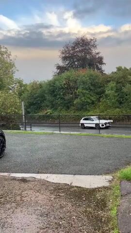 The video shows an outdoor scene with a white car parked on a wet road. The car is facing to the right. Behind the car is a dense line of green trees and bushes. A large, dark-colored tree with reddish-brown leaves is visible above the green foliage. A black metal fence runs along the edge of the road. The sky is overcast with clouds, and there are patches of blue sky visible. The foreground of the video shows a gravel area with a wet patch. The video is shot from a low angle, looking slightly upwards.