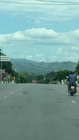 This media file captures a view from a vehicle traveling on a paved road in Lao Bảo Commune, Vietnam. The scene is dominated by a wide, asphalt road with yellow lane markings. In the foreground, a motorcycle with a rider is visible on the right side of the road. Further ahead, a red semi-trailer truck is passing a black car, and another motorcycle is approaching from the opposite direction.

The setting is rural, with lush green trees lining both sides of the road. Beyond the trees, rolling hills covered in vegetation stretch towards the horizon, where a hazy mountain range can be seen. A cell tower stands prominently on one of the hills. The sky is a bright blue with scattered, fluffy white clouds, suggesting a sunny day. A streetlamp is visible on the left side of the road, and power lines run along the right.

There are a few signs visible. On the left, there are some white signs with text, but they are too far away to be legible. On the right side of the road, near the approaching motorcycle, there is a yellow diamond-shaped sign with black symbols, likely indicating a warning.

The overall impression is one of travel through a scenic, developing area, with everyday transportation activities taking place. The time of day appears to be midday, given the bright sunlight and clear sky. There are no strong emotional cues conveyed in the media.