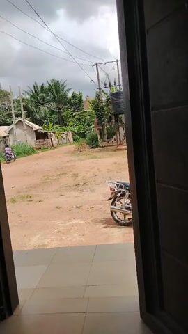 The video, filmed from inside a doorway with a tiled floor, looks out onto a rural or semi-urban landscape in Akwa Etiti, Nigeria. The scene features an unpaved, reddish-brown dirt area or road as the foreground and midground. In the immediate right foreground, a motorbike is parked, with its rear wheel and luggage rack visible.

In the distance, on the left side of the frame, simple, likely residential structures with unpainted walls and basic roofing are visible amongst lush green vegetation, including palm trees and what appear to be banana plants. Power poles with overhead wires crisscross the sky, and a large black water tank on a stand is visible on the right side, behind the parked motorbike.

The sky is heavily overcast with grey clouds, suggesting either an approaching storm or an overcast day. Briefly, a person on a motorbike is seen driving away into the distance on the dirt road before disappearing from view. The overall impression is a quiet, natural setting with basic infrastructure.